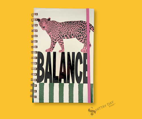 Notebook with "Balance" text in bold black, sitting on thick green vertical lines. A large pink spotted wild feline is walking on the top of the letters. The notebook is wire bound and shows a pink elastic band closure. The image is featured on an yellow background.