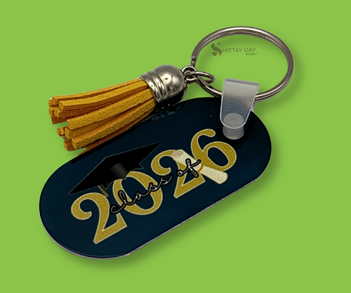 Graduation-themed Navy Blue oval shaped keychain with "Class of 2026" gold text and a grad cap and rolled degree image. The Keychain has a yellow tassel. Image is featured on a bright green background.