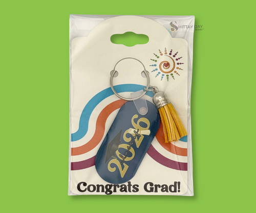 Graduation-themed navy blue keychain with "Class of 2026" design text and a black grad cap plus rolled degree rolled. The keychain also has a yellow tassel. The keychain is in clear packaging with colorful design merchandising back card, featured on a bright green background.