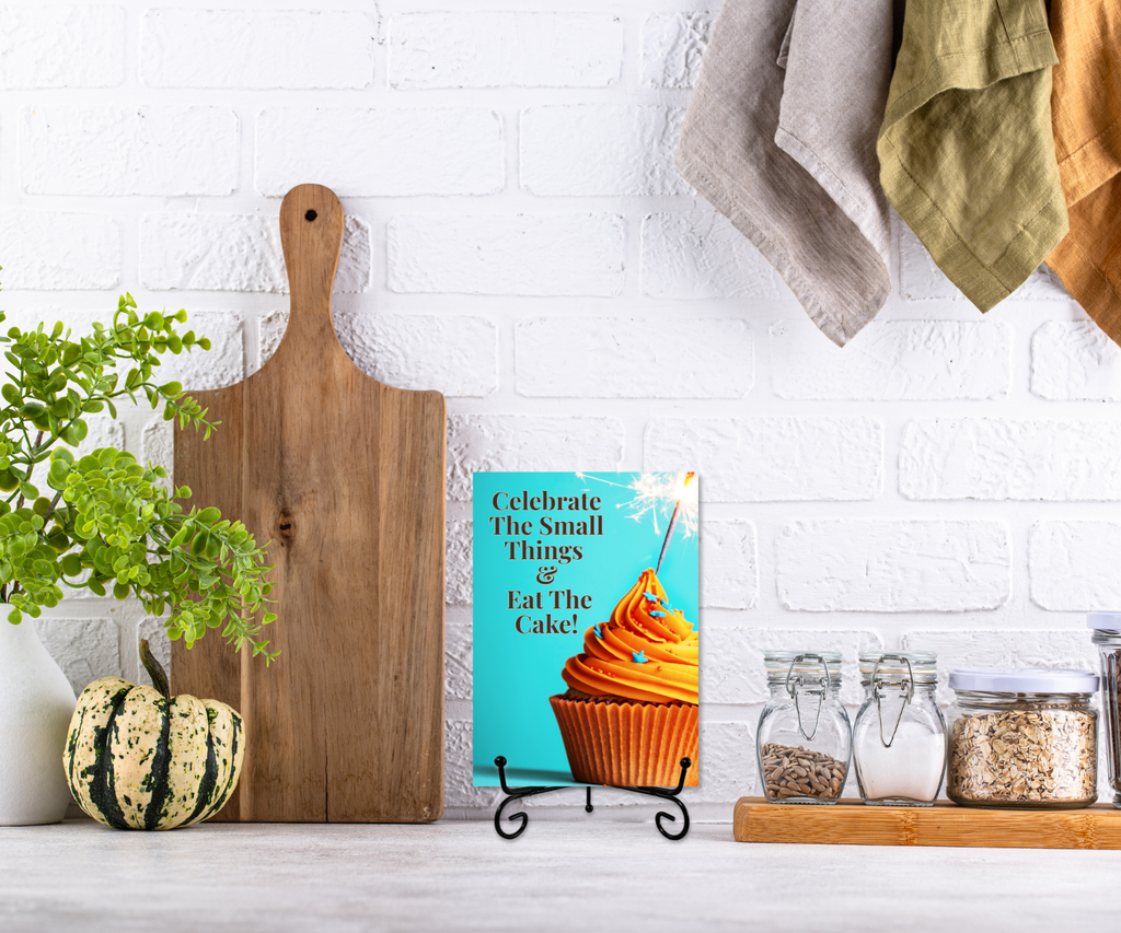 An aluminum keepsake greeting card with a cupcake and the text "Celebrate the small things & eat the cake" on a metal stand displayed on a kitchen counter. 