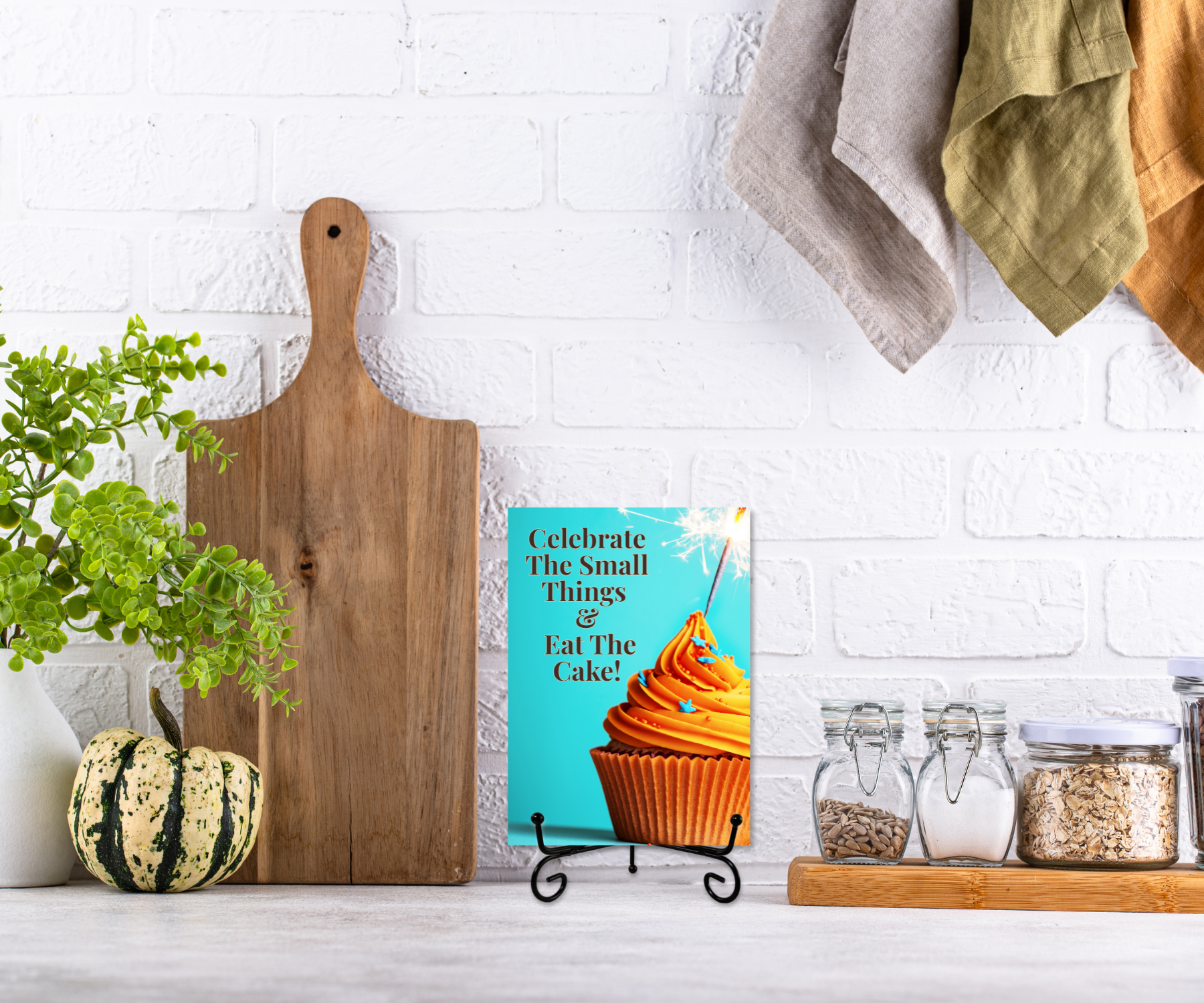 An aluminum keepsake greeting card with a cupcake and the text "Celebrate the small things & eat the cake" on a metal stand displayed on a kitchen counter. 
