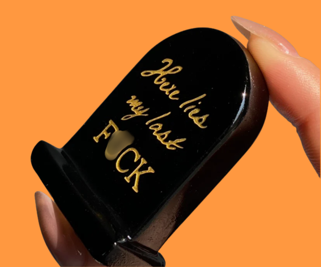 Natural crystal “Here lies my last F*CK” tombstone held between fingertips to show its small size.
