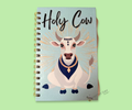 A wire bound notebook with a cow wearing a collar and necklace illustration and 'Holy Cow' text on a light blue background with gold starbursts and white fleck dots. Featured on a green background