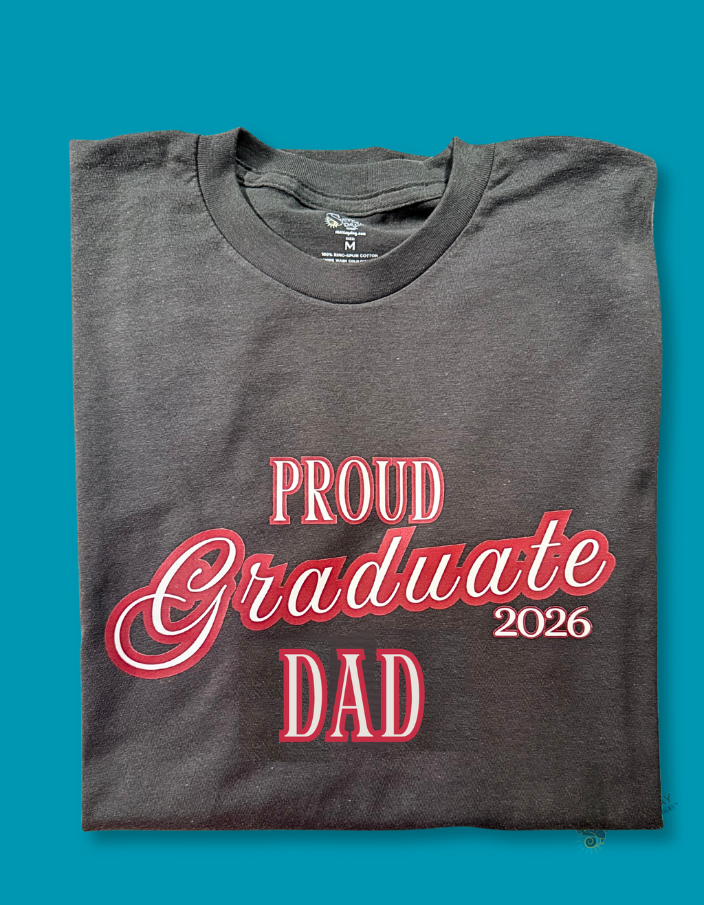 Gray color folded tshirt with a close up of the design; 'Proud Graduate Dad 2026'  maroon outlined white l text. Merchandised on a blue background.