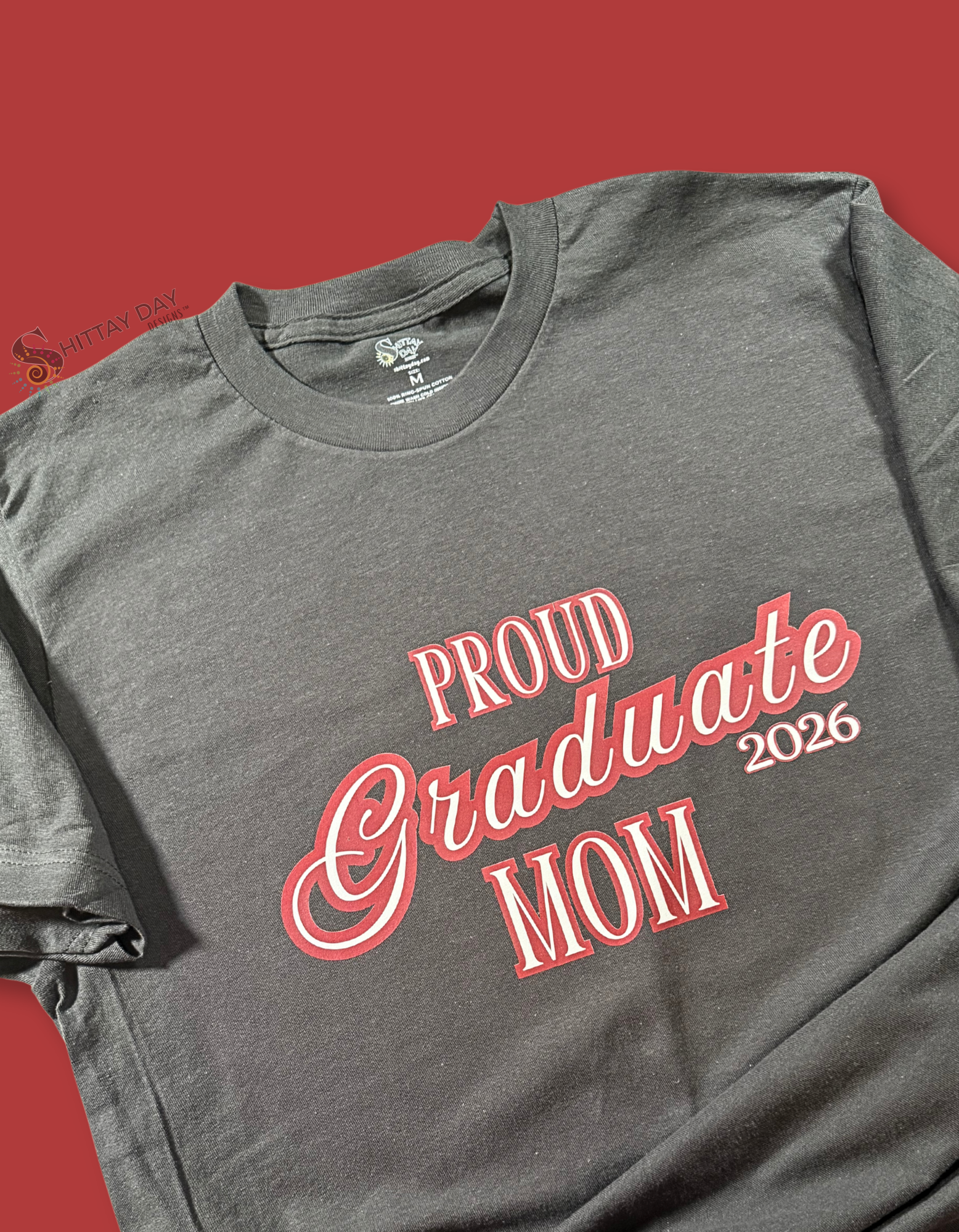 Gray color folded tshirt with a close up of the design; 'Proud Graduate Mom 2026'  maroon outlined white l text. Merchandised on a maroon background.