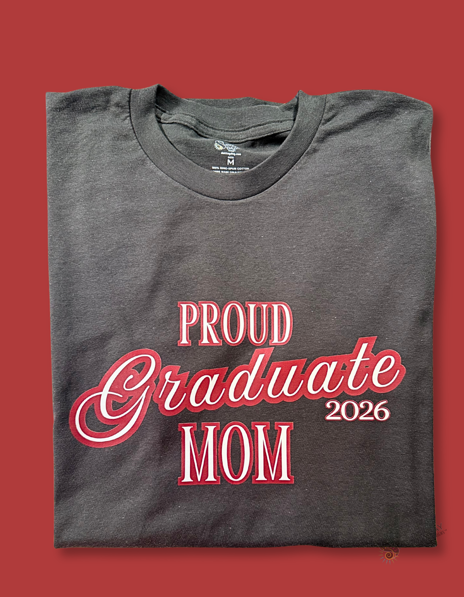 Gray color folded tshirt with a close up of the design; 'Proud Graduate Mom 2026'  maroon outlined white l text. Merchandised on a blue background.