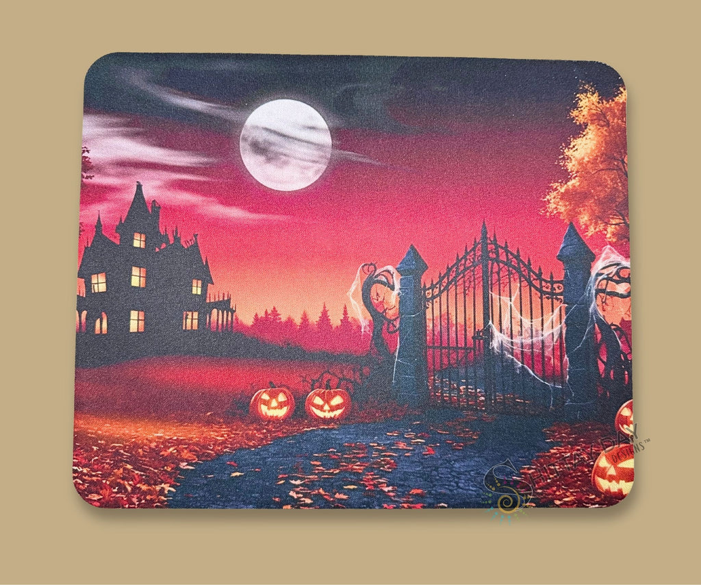 A mouse pad with a Halloween-themed illustration, featuring a haunted house, full moon, pumpkins, and a gate, shown with a white  mouse on top.