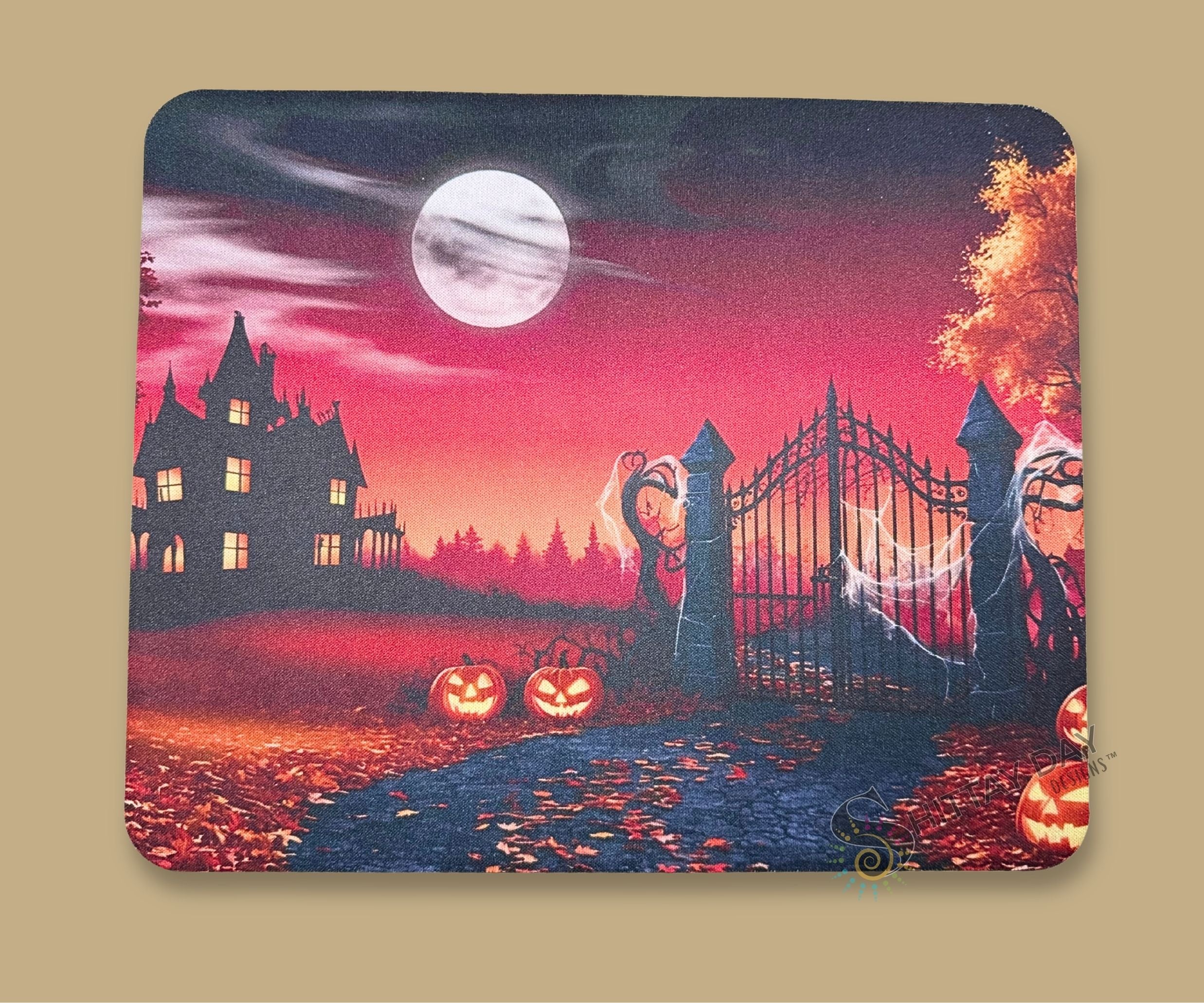 A mouse pad with a Halloween-themed illustration, featuring a haunted house, full moon, pumpkins, and a gate, shown with a white  mouse on top.
