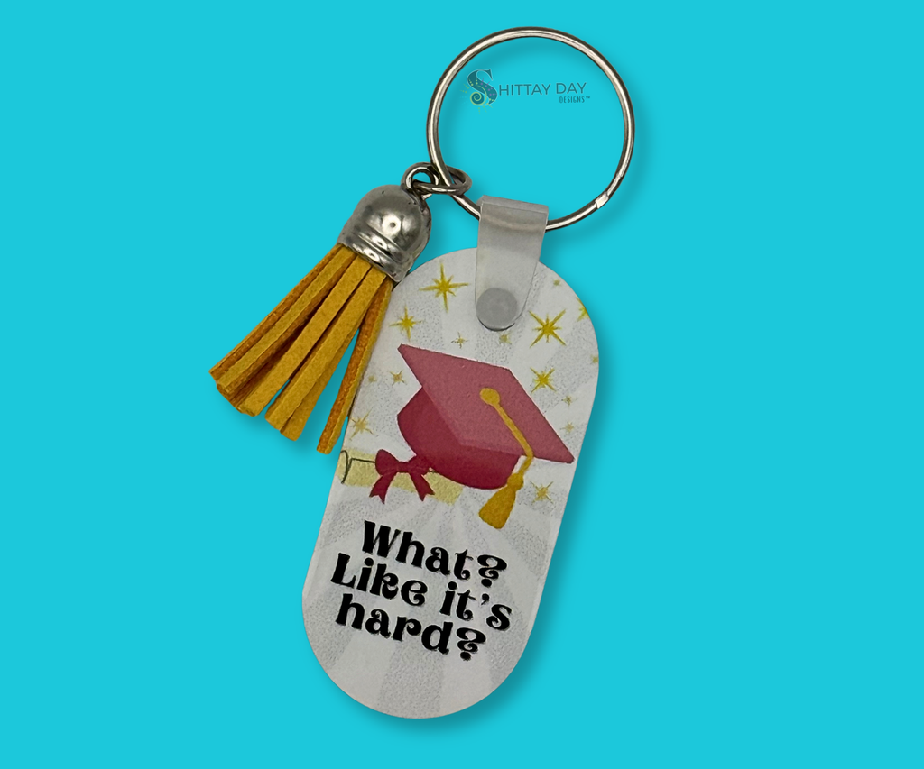 Graduation-themed keychain with "What? Like it's Hard?"  text and a pink grad cap, degree rolled with pink ribbon, and yellow starbursts. Keychain has a yellow tassel. Image is featured on a blue background.