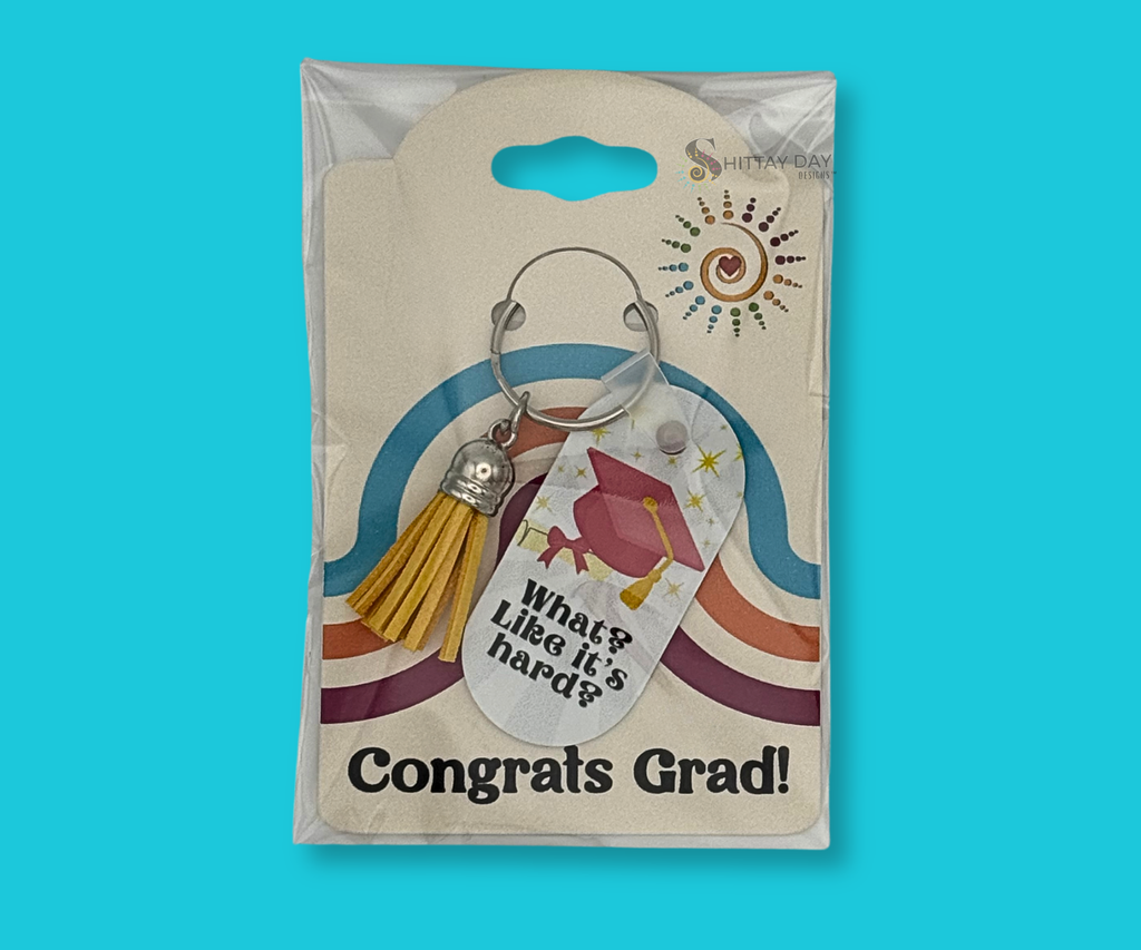 Graduation-themed keychain with "What? Like it's Hard? text and a pink grad cap, degree rolled with pink ribbon, and yellow starbursts keychain with a yellow tassel, in clear packaging with colorful design merchandising back card, featured on a blue background.