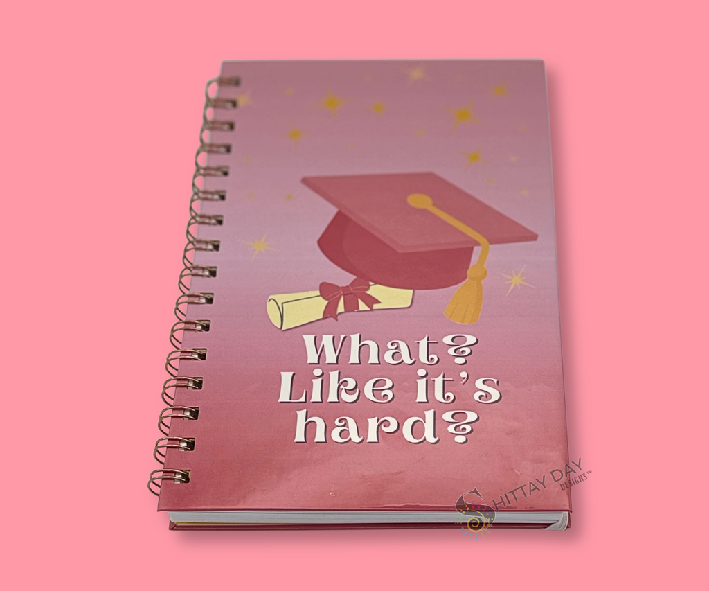 Wire bound notebook with graduation design featuring a pink grad cap and rolled diploma with a pink bow, pink gradient background with yellow stars and text "what? like it's hard? featured on a pink background