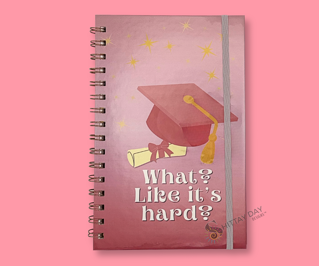 Wire bound notebook with graduation design featuring a pink grad cap and rolled diploma with a pink bow, pink gradient background with yellow stars and text "what? like it's hard? featured on a pink background