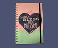 A wire bound notebook with heart design and text "well bless your little heart" on a pink and green background with floral sketches. features on a blue background